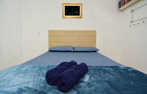 A bedroom with a wooden headboard, two pillows, a blue bedspread, and rolled towels on the bed. - Casa Aconchegante na Lagoa da Cigana (Laguna)