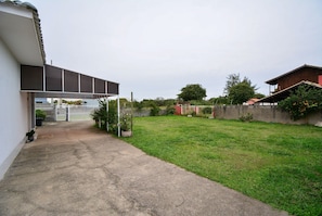 A residential property with a paved driveway, a grassy yard, and a red building in the background. - Casa Aconchegante na Lagoa da Cigana (Laguna)