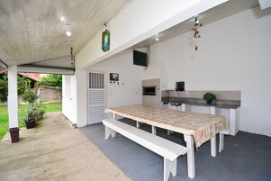 Uma área de refeições coberta ao ar livre, com mesa e bancos, um pequeno espaço de cozinha e uma porta que dá para um alpendre coberto.