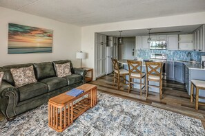 Ein Wohnzimmer mit Sofa, Couchtisch und Sesseln, eine Küche mit Essbereich sowie ein Gemälde an der Wand.