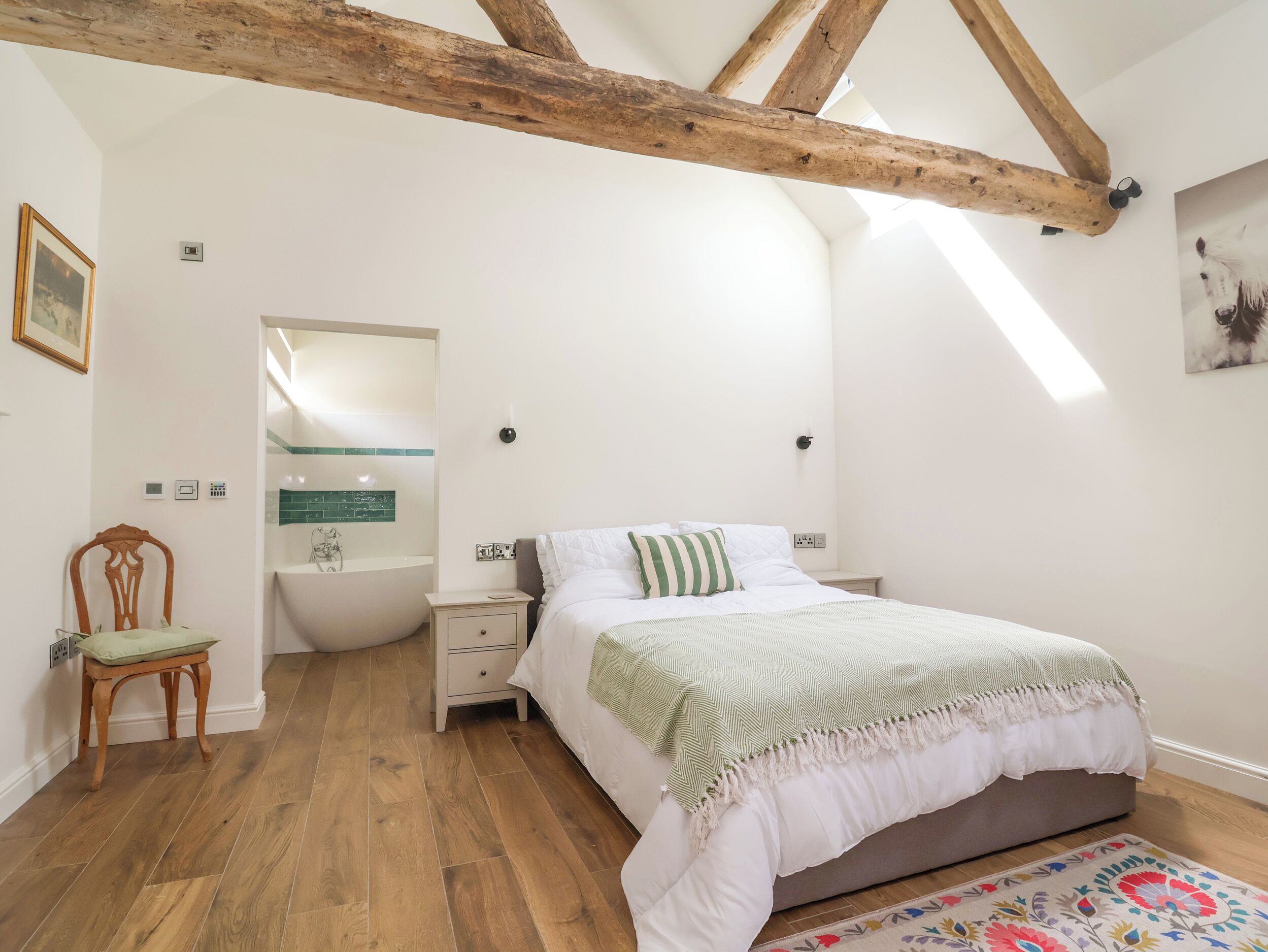A bedroom with a wooden ceiling, a bed with white bedding, a bedside table, a chair, and a bathroom with a bathtub.