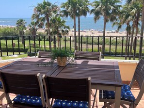 Ein Holztisch mit Stühlen und einem Blumentopf auf einem Balkon mit Blick auf einen Strand, an dem Palmen stehen, und einem klaren Himmel.