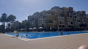 Et poolområde med liggestole, en poolterrasse og en flereetagers bygning i baggrunden.