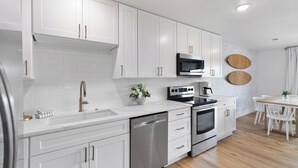 A modern kitchen with white cabinetry, stainless steel appliances, and a wooden dining area. - My Blue Heaven 30A Getaway - Beach Access, Pools & Prime 30A Location (Santa Rosa Beach)