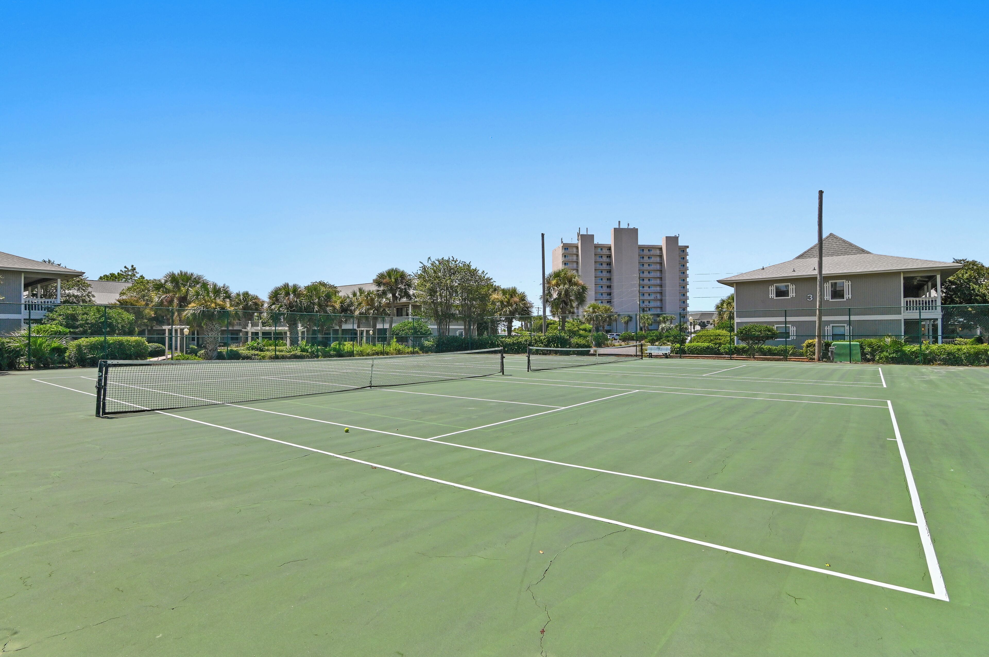 Una cancha de tenis con red, rodeada de vegetación y edificios.