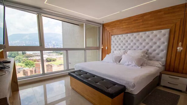 A bedroom with a large window, a bed with a tufted headboard, a wooden bench, and a nightstand. - Amplio ático con vistas, jacuzzi a 15 min de Provenza (Itagüí)