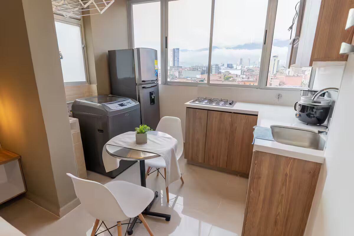 Una cocina compacta con refrigerador, una pequeña mesa con sillas, un ventanal con vista a la ciudad y un lavamanos.