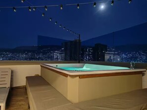 Eine Dachterrasse mit Whirlpool, Sitzgelegenheit und Blick auf die nächtliche Stadt.