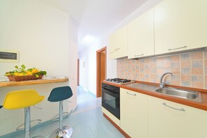 A compact kitchen with a gas stove, oven, and sink. There are two chairs and a small table with a basket of lemons. - Armonia (Barano D'ischia)