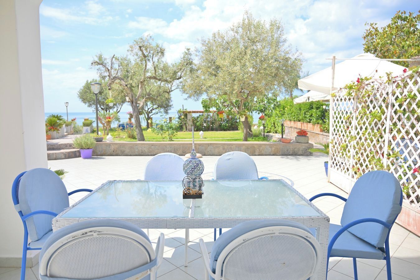 A glass-top table with four chairs, a decorative lamp, and a view of the sea and greenery.