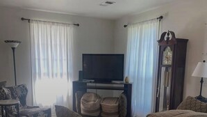 Un soggiorno con televisione su un mobile, un divano, una poltrona con motivo decorativo, una lampada e una sveglia.