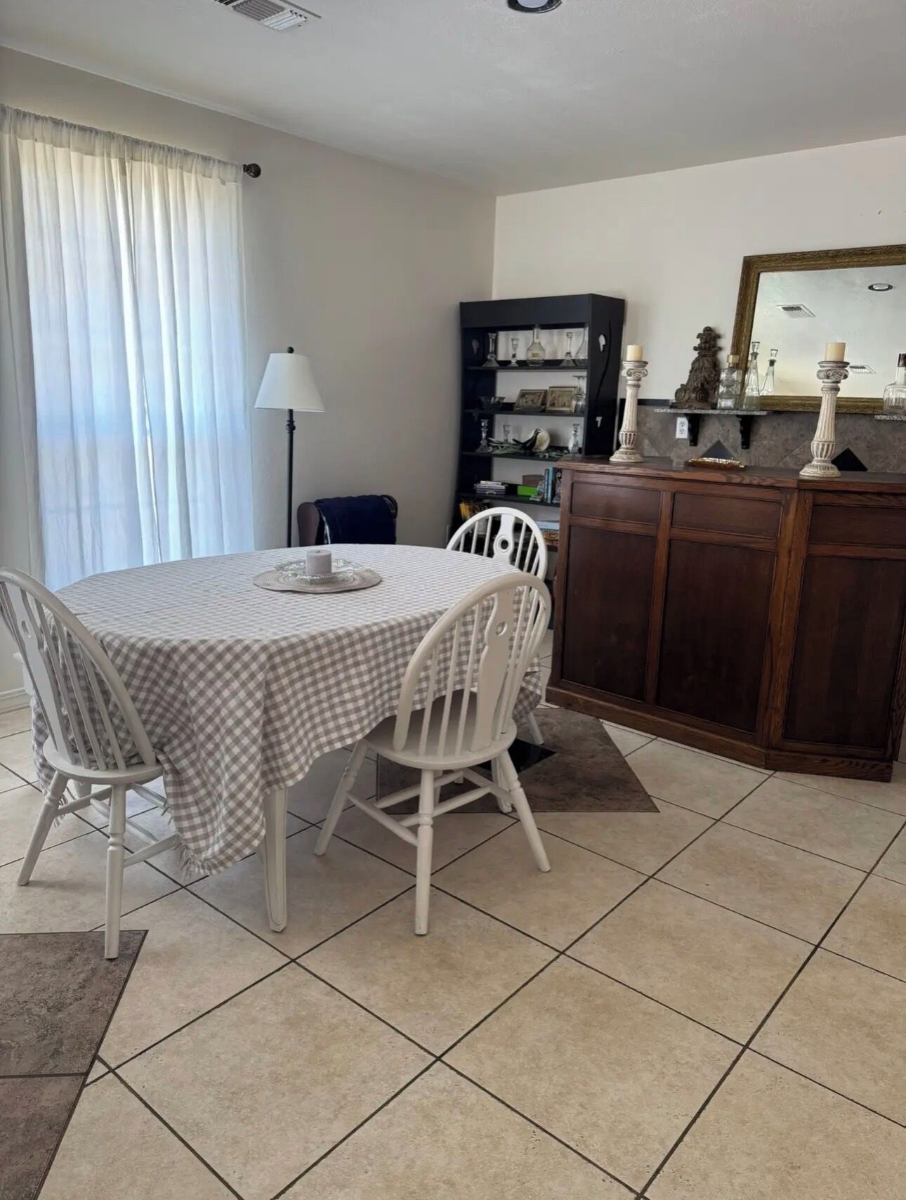 Un coin repas avec une table ronde, des chaises blanches, une nappe à carreaux, une lampe, une bibliothèque et un meuble contenant des objets décoratifs.