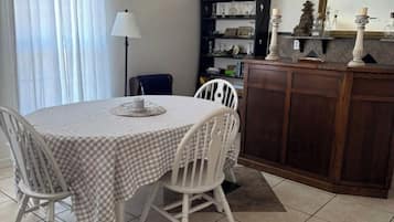 Un coin repas avec une table ronde, des chaises blanches, une nappe à carreaux, une lampe, une bibliothèque et un meuble contenant des objets décoratifs.