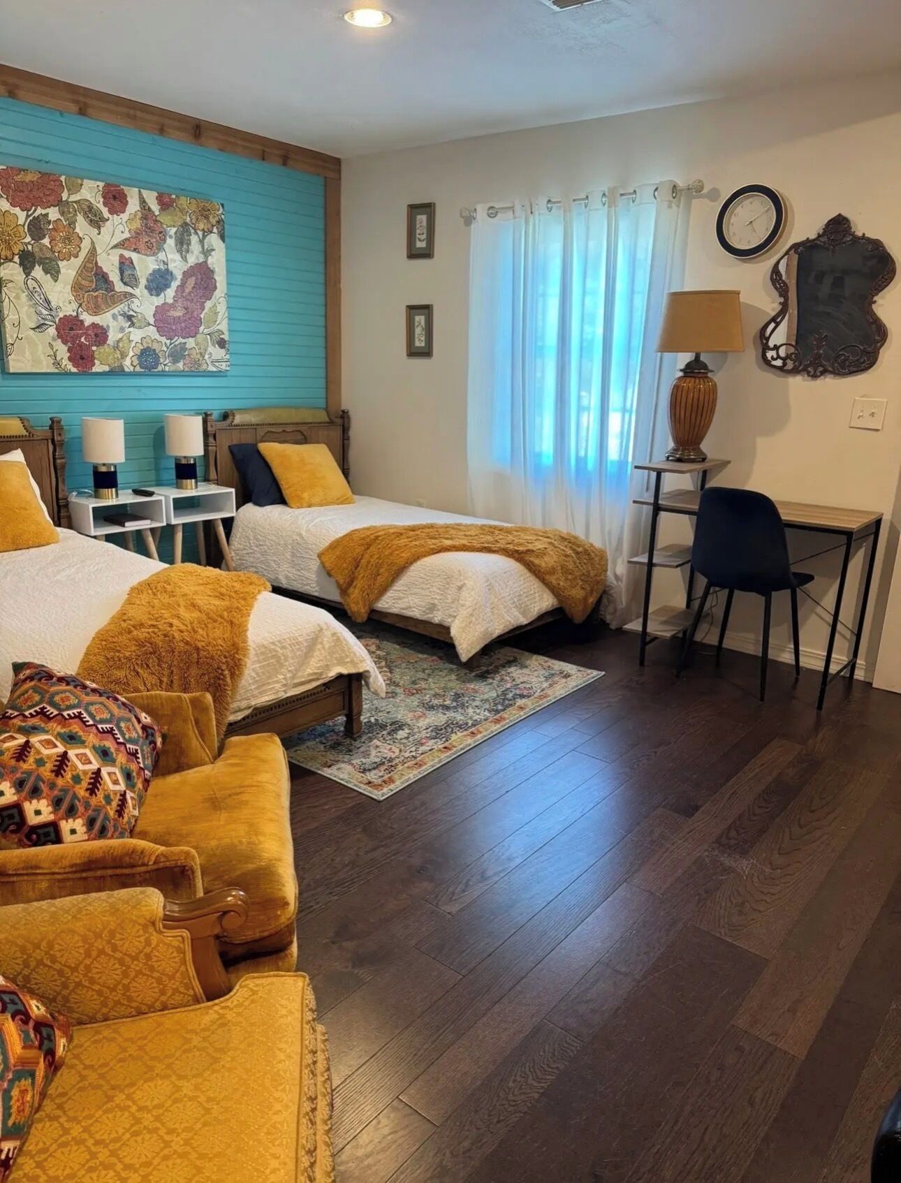 Une chambre d’hôtel avec deux lits, un bureau, une chaise, une horloge et une œuvre d’art florale accrochée au mur.