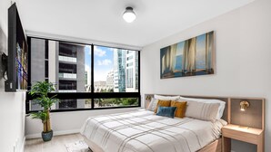 A modern bedroom with a large window, a bed with a headboard, a nightstand, a potted plant, and a painting on the wall. - Luxury 1BR Downtown Miami I Free Parking + Pool (Miami)