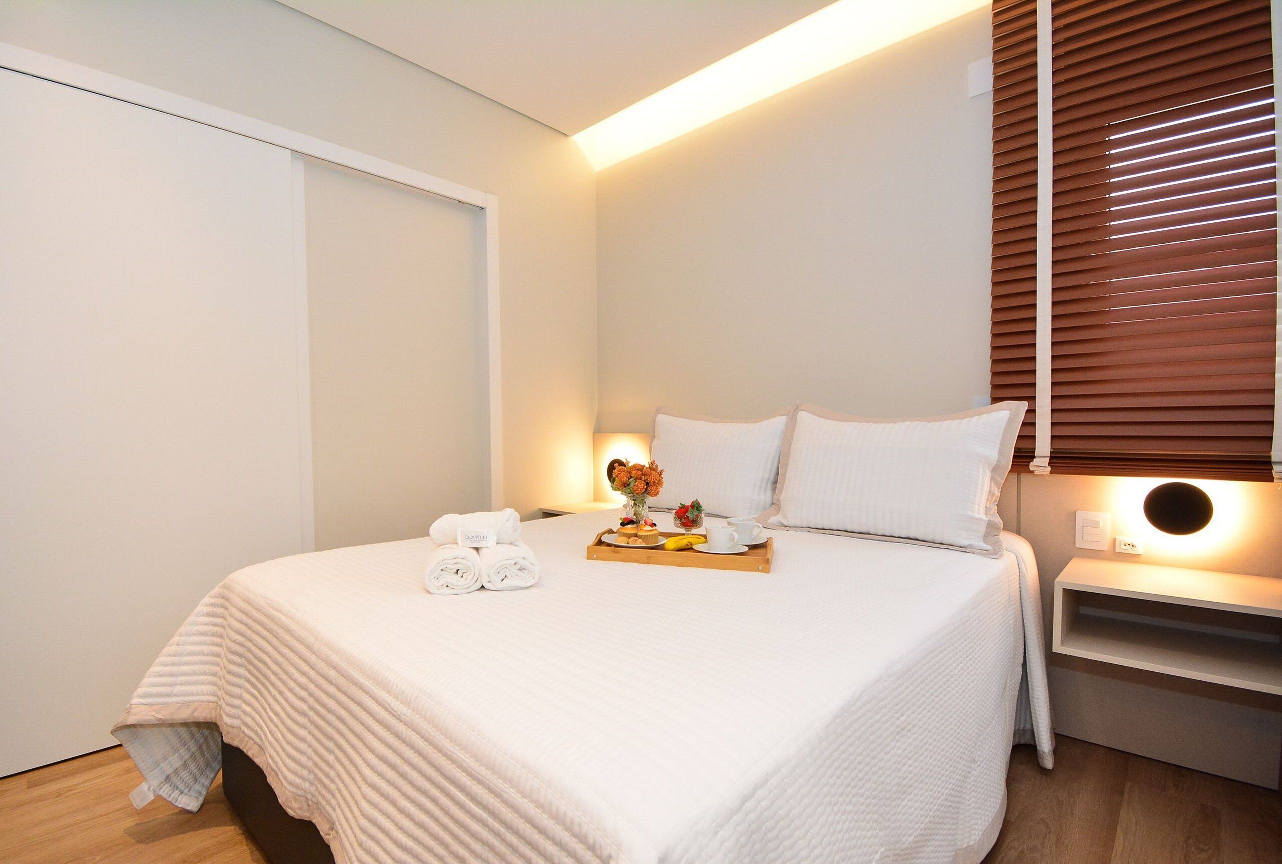 A neatly made bed with a tray of breakfast items, a bedside table with a lamp, and a window with blinds.