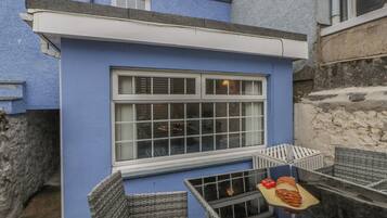 A blue building with a large window, a small balcony with wicker furniture, and a wooden table with bread and tomatoes.