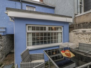 Un edificio azul con un amplio ventanal, un balcón pequeño con mobiliario de mimbre y una mesa de madera con pan y tomates.
