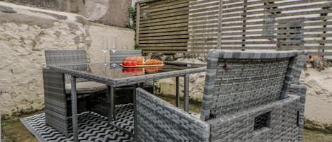 Un espacio de estar al aire libre con muebles de mimbre, una mesa y sillas de vidrio, un cerco de madera y un muro de piedra al fondo.