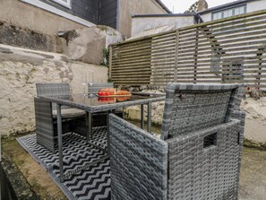 Un espacio de estar al aire libre con muebles de mimbre, una mesa y sillas de vidrio, un cerco de madera y un muro de piedra al fondo.