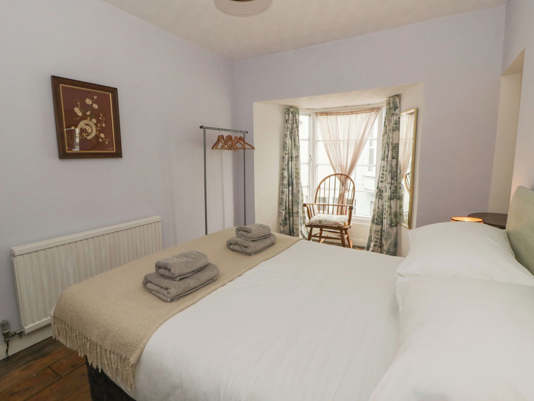 Un dormitorio con cama, una silla, un radiador, un cuadro en la pared y una ventana con cortinas.