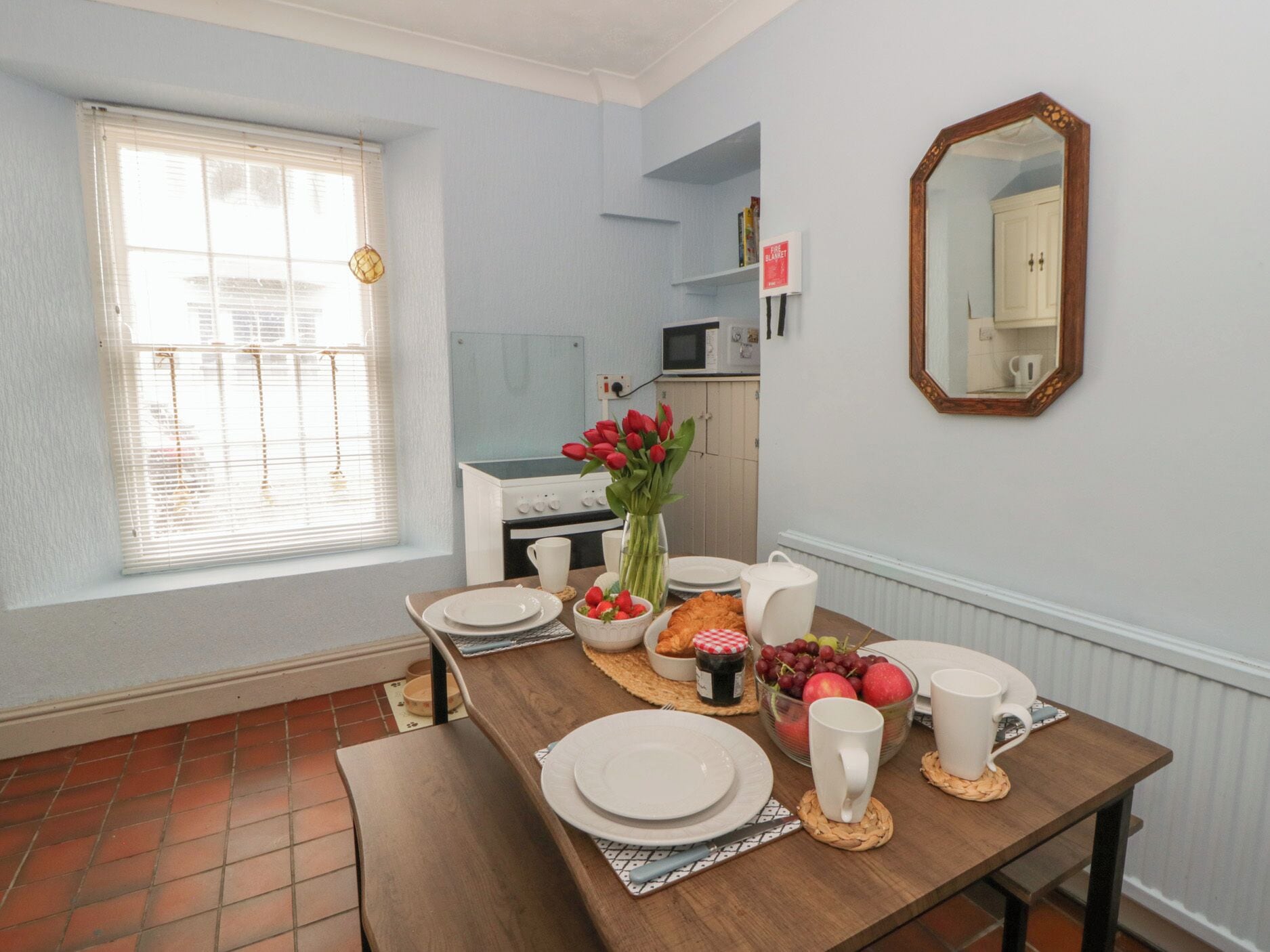 Un comedor con una mesa dispuesta para cuatro personas, un jarrón de tulipanes rojos y una cocina al fondo.