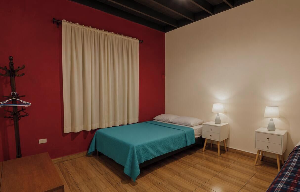 Un dormitorio con una cama, dos mesitas de noche con lámparas, suelo de madera, pared roja y una ventana con cortinas.