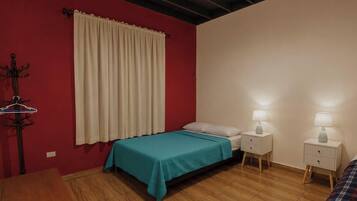 Un dormitorio con una cama, dos mesitas de noche con lámparas, suelo de madera, pared roja y una ventana con cortinas.