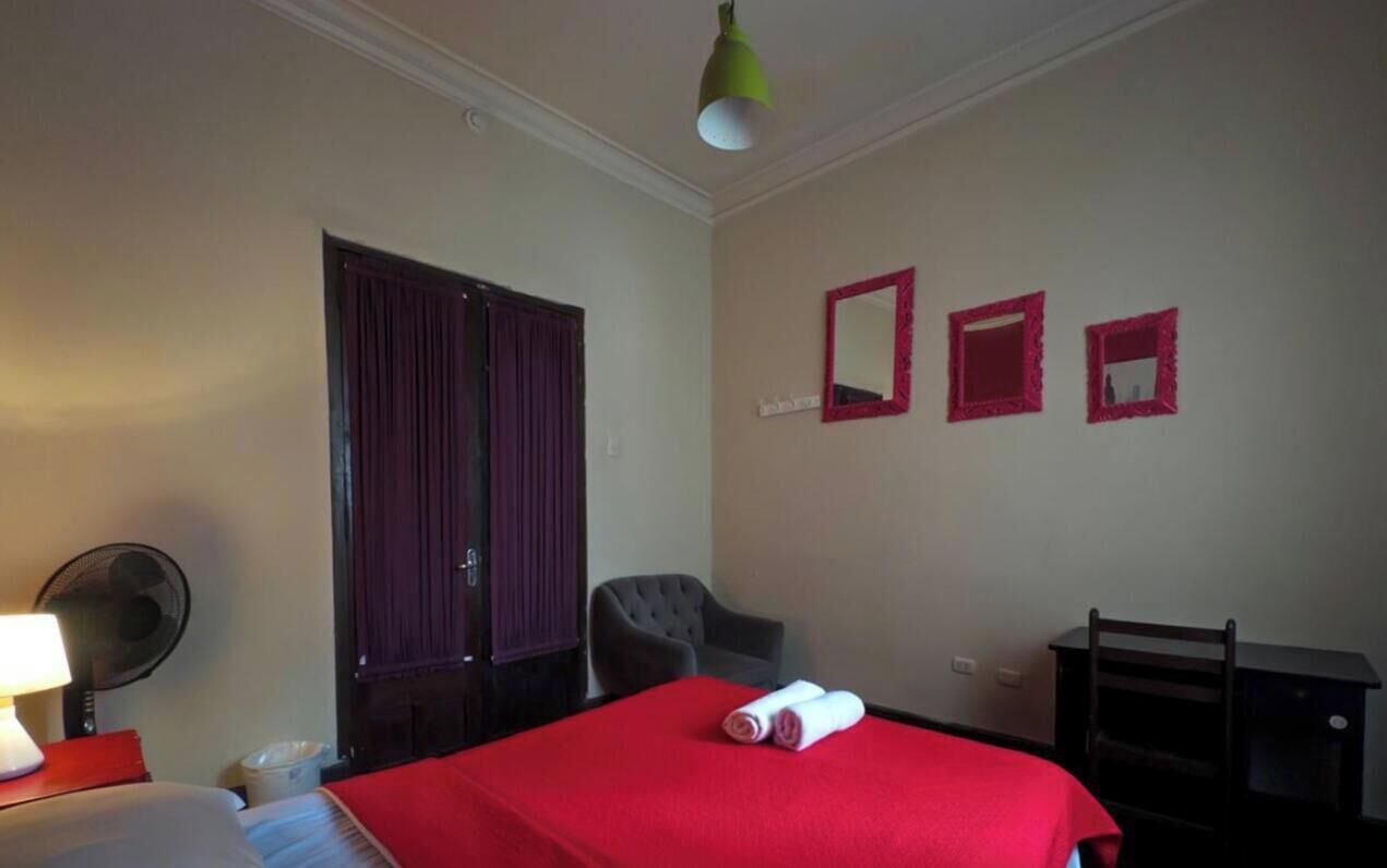 A hotel room with a bed, a chair, a desk, a lamp, and a fan.