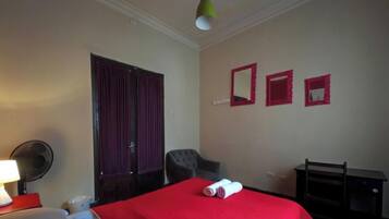 A hotel room with a bed, a chair, a desk, a lamp, and a fan.