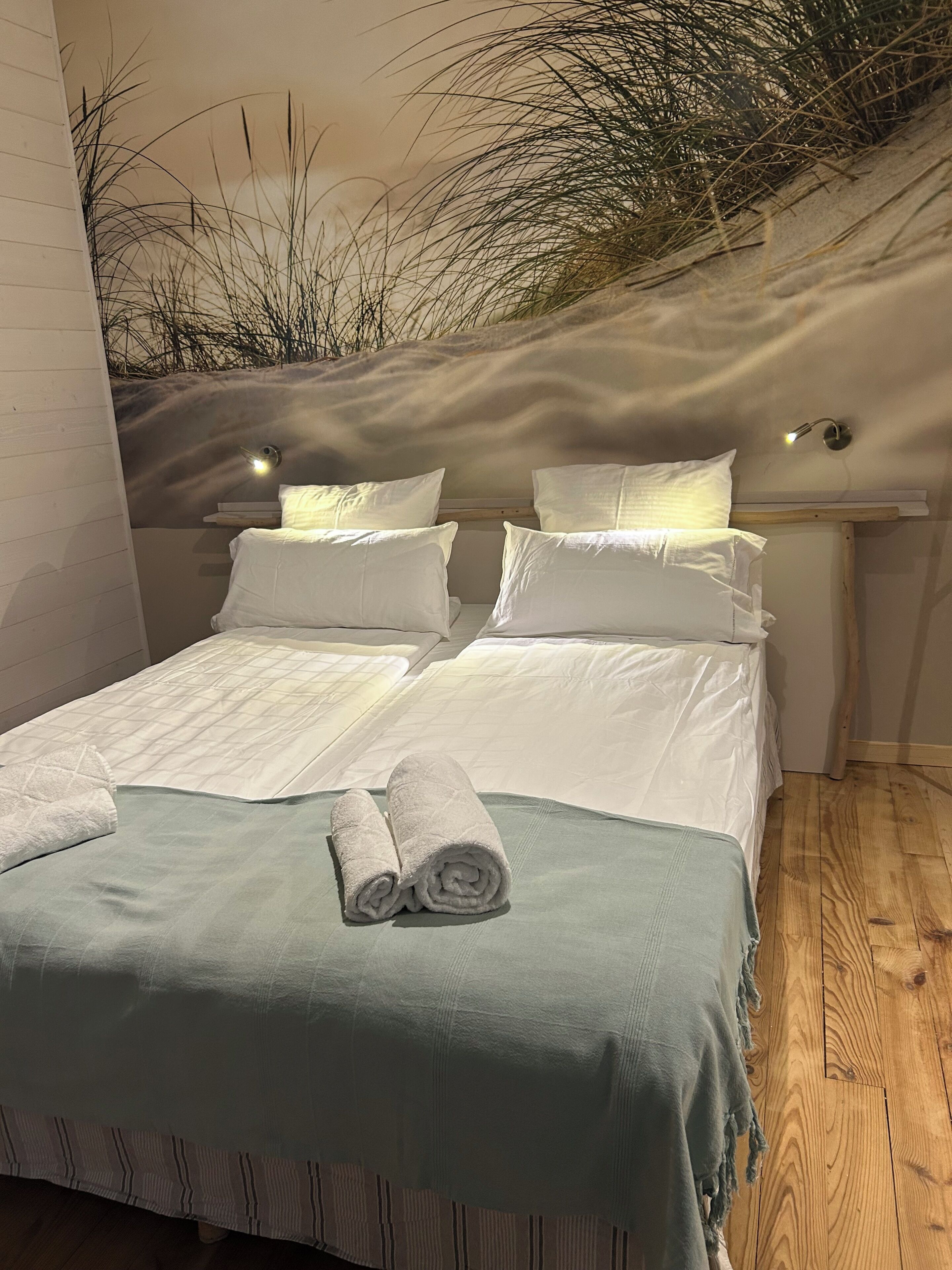 Una cama doble con sábanas blancas, una manta verde y toallas enrolladas sobre la cama.
