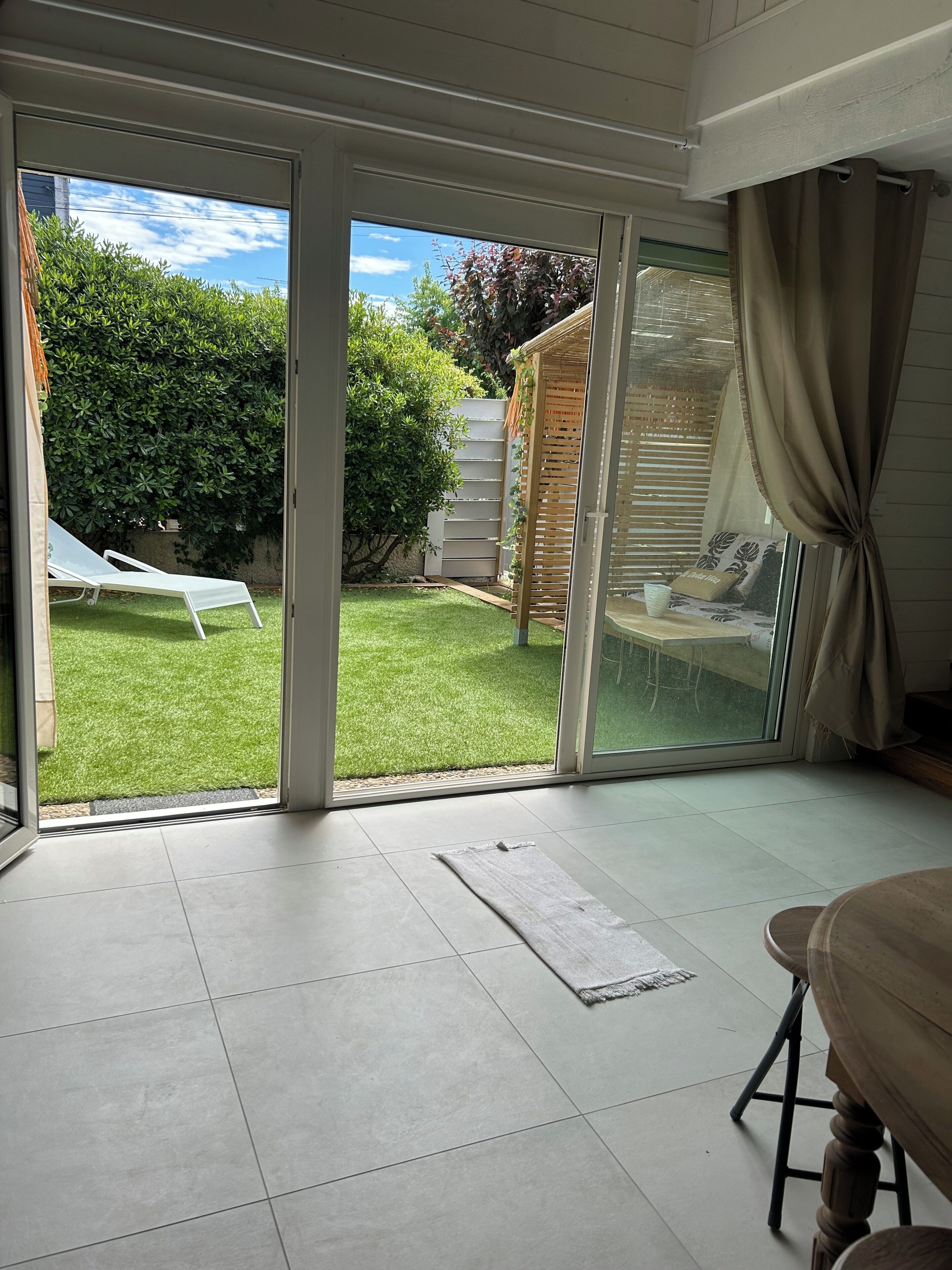 A room with a sliding glass door leading to a backyard with a lawn, a wooden pergola, and a white lounge chair.