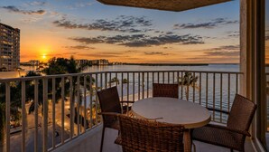 Ein Balkon mit Korbstühlen und einem Tisch, der Blick geht auf einen Sonnenuntergang über dem Wasser, im Hintergrund sind Gebäude zu sehen.