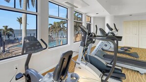 Ein Fitnessraum mit Laufbändern, Crosstrainern und Rudergeräten, mit großen Fenstern mit Blick auf einen Pool und Palmen.