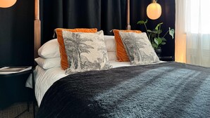 A bed with a dark grey blanket, two pillows with tropical prints, and a nightstand with a lamp. - The Tenter Ground  Liverpool Street (London)