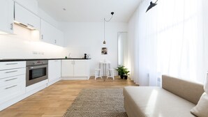 Moderni keittiö, jossa on valkoiset kaapit, ruokailutila, jossa on valkoinen pöytä ja tuolit, sekä olohuone, jossa on beigenvärinen sohva.
