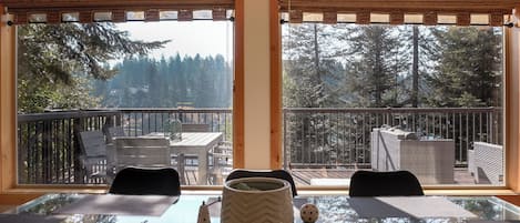 A dining area with a glass table, two chairs, and a view of a balcony with outdoor furniture and trees.