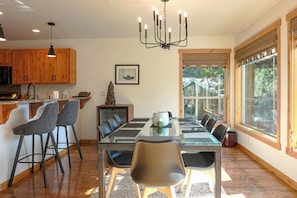 A modern kitchen with wooden cabinets, a dining area with a glass table, and a large window with a view of trees.