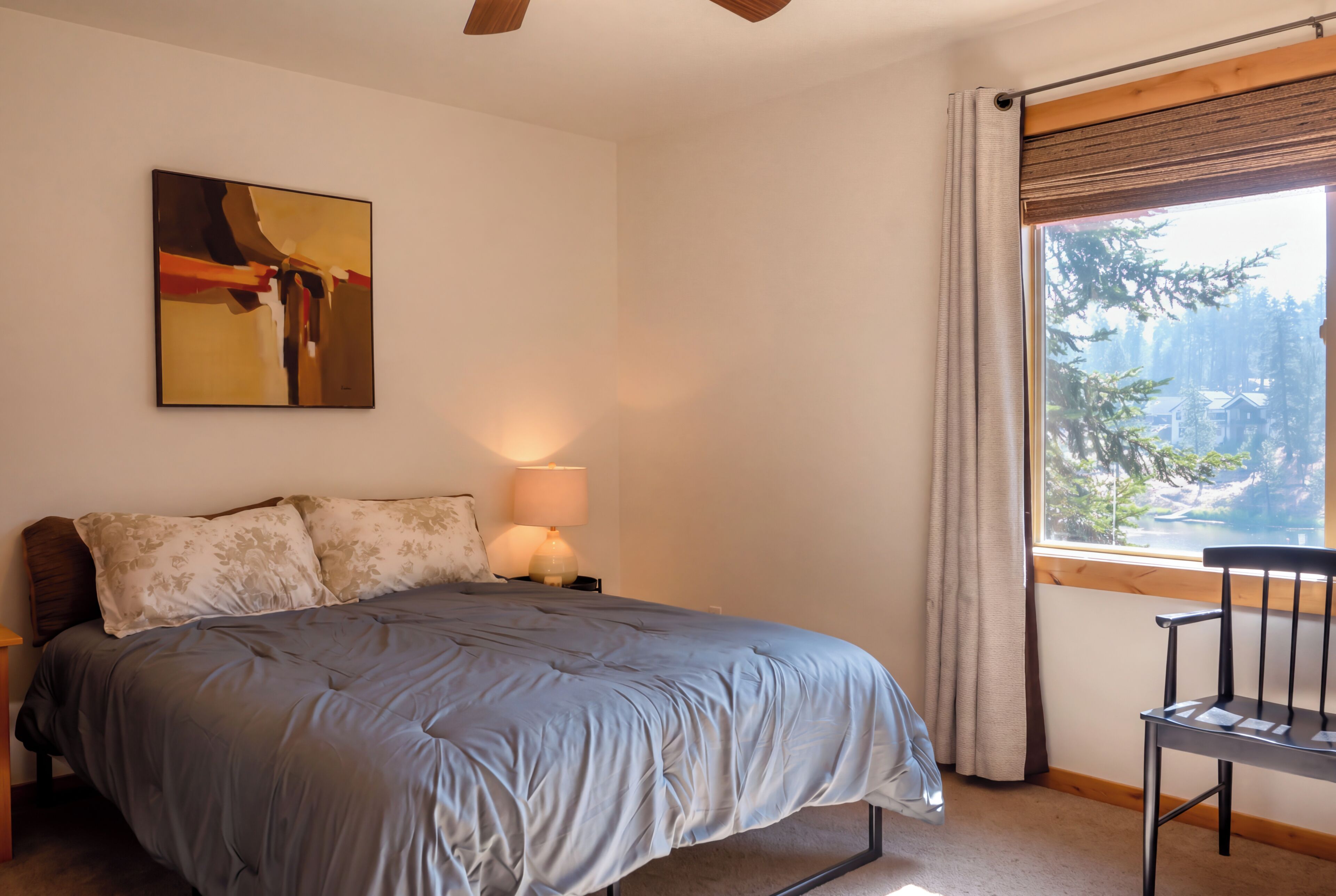 A bedroom with a bed, a nightstand, a lamp, a chair, a window with a view of trees and a lake, and a painting on the wall.