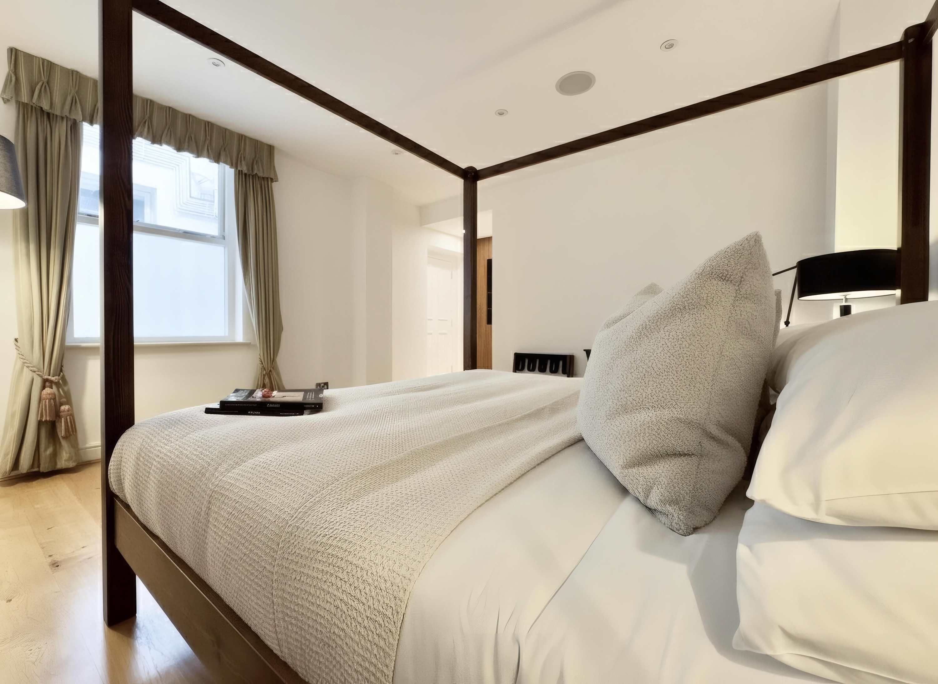 A bedroom with a four-poster bed, a bedside table, a lamp, and a window with curtains.