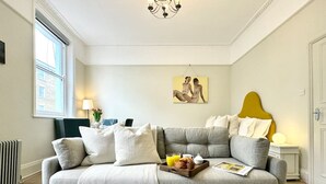 A living room with a grey sofa, a coffee table with breakfast items, a painting on the wall, and a radiator. - Chic Marylebone Village  Premium Mattress   55   TV (London)