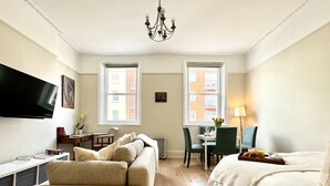 A hotel room with a sofa, a bed, a desk with a chair, a TV, and two windows with a view of buildings. - Chic Marylebone Village  Premium Mattress   55   TV (London)