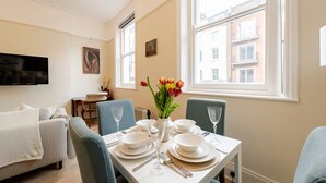 A dining area with a table set for four, a sofa, a TV, and a window with a view of buildings. - Chic Marylebone Village  Premium Mattress   55   TV (London)