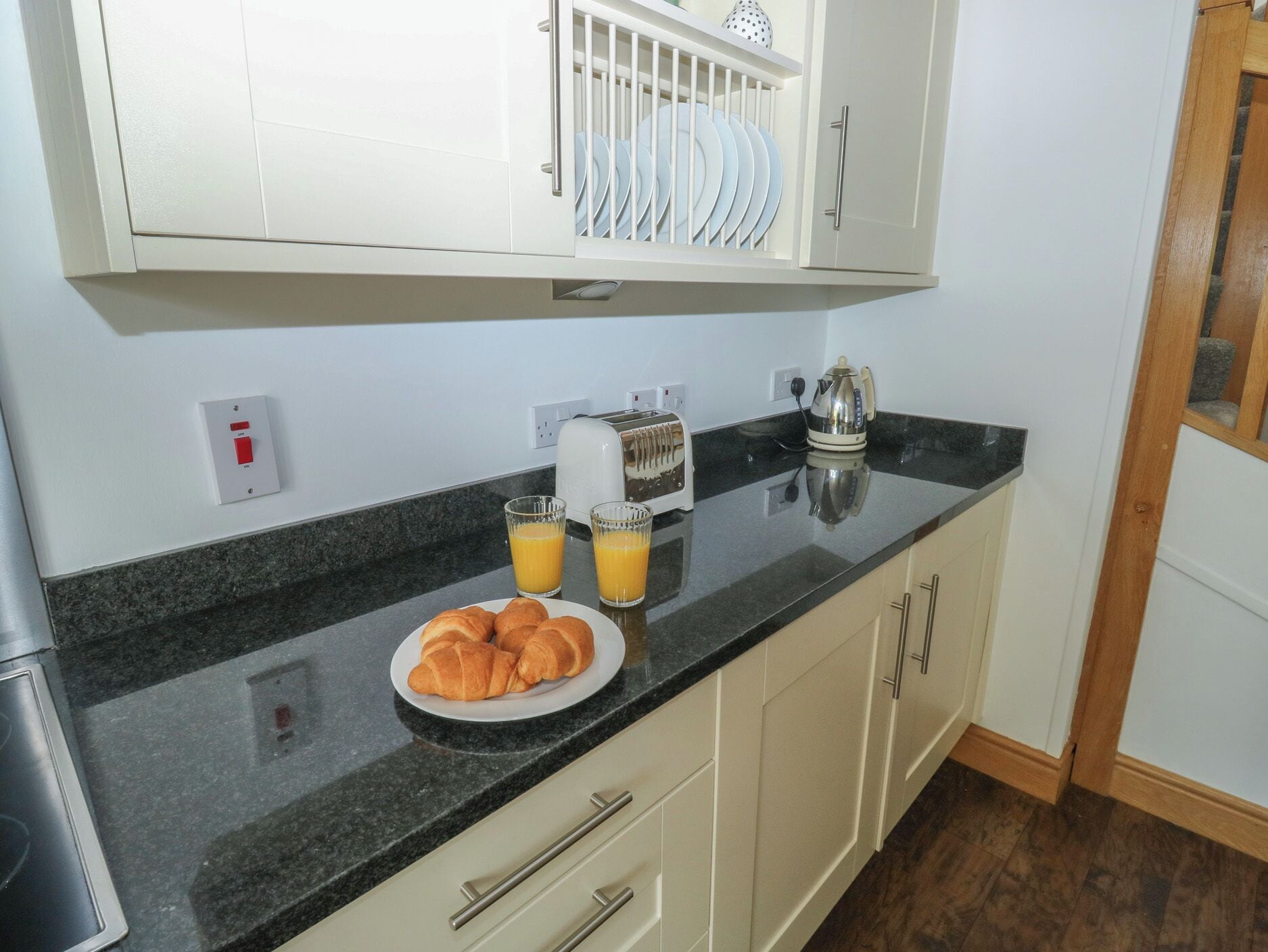 Uma cozinha com bancada preta, um prato de croissants, dois copos de suco de laranja, uma torradeira e uma chaleira.