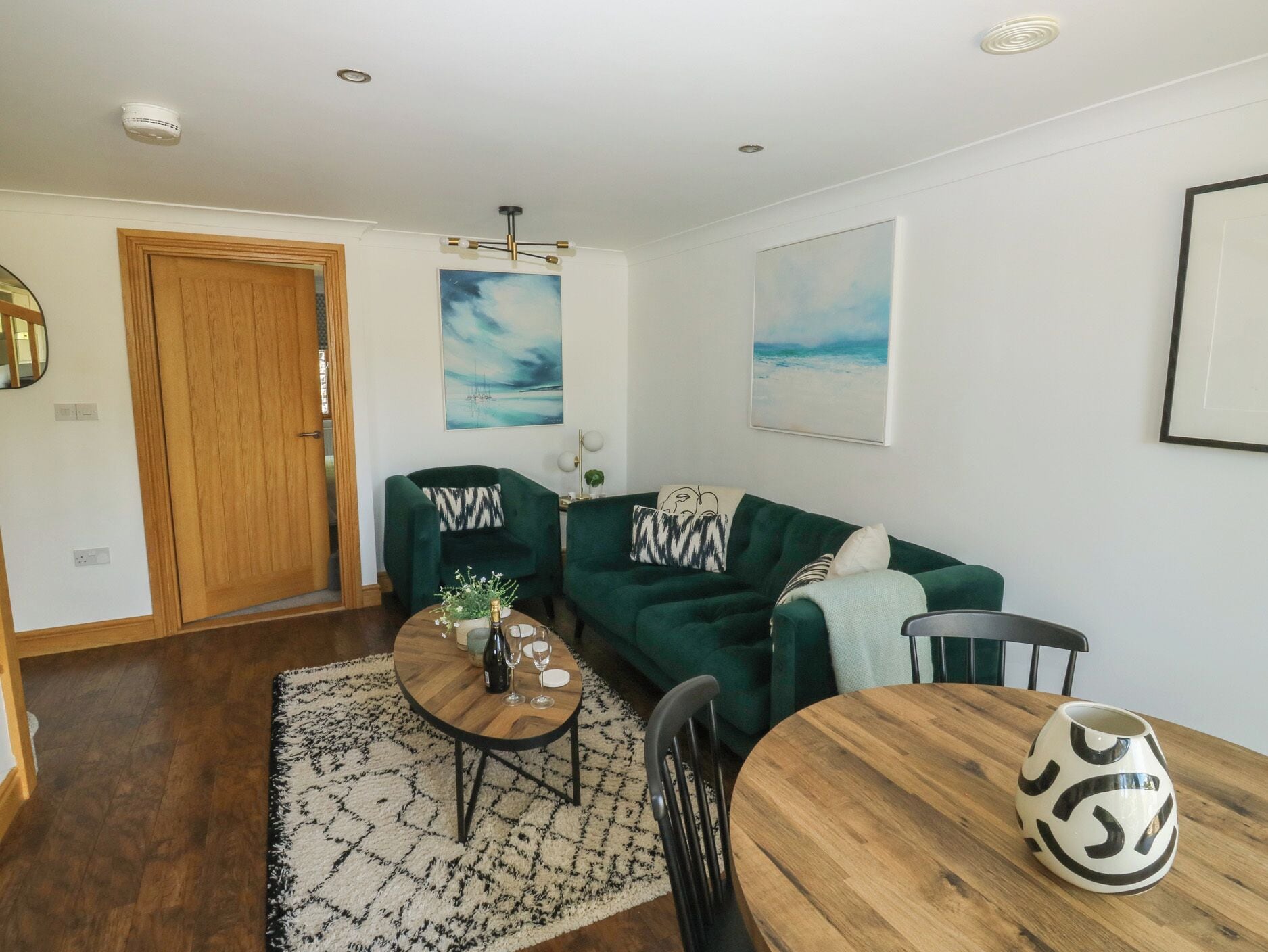 A living room with a green sofa, a wooden coffee table, and a dining table with chairs. There are two paintings on the wall, a wooden door, and a ceiling light fixture.