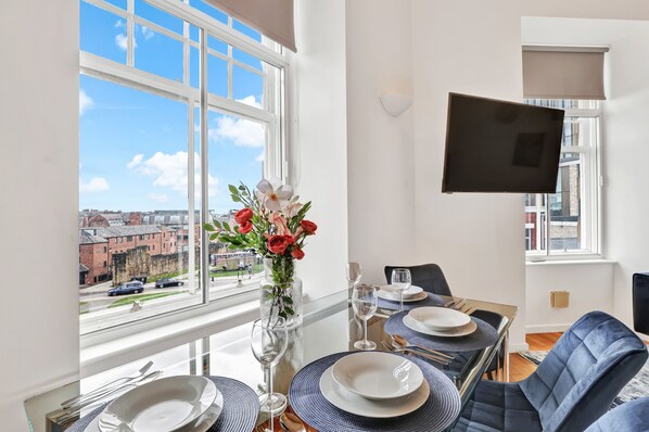 Ein Essbereich mit einem Tisch für vier Personen, einer Blumenvase und Blick auf die Stadt durch große Fenster.