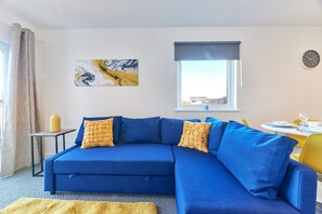 A modern living room with a blue sectional sofa, a small side table, a yellow throw blanket, a large abstract painting, a window with a view, and a dining area with yellow chairs. - Mindrum in North Shields (North Shields)