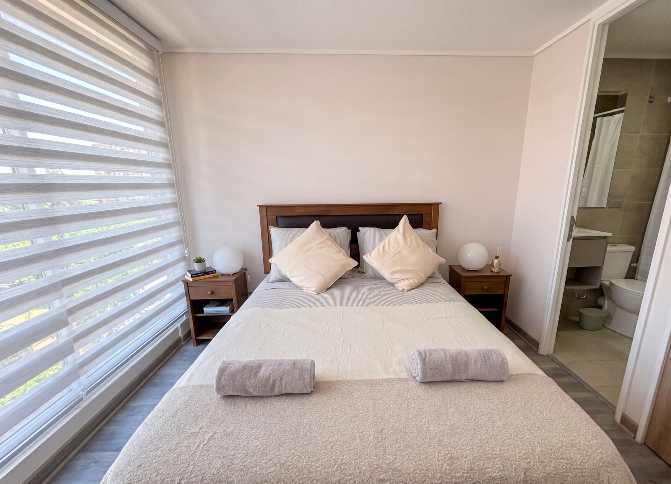 Een slaapkamer met een groot bed, twee nachtkastjes, een raam met jaloezieën en uitzicht op de badkamer.