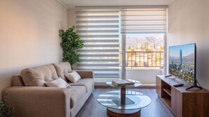 Una sala de estar moderna con un sofá, una mesa de centro de vidrio, un televisor sobre un mueble de madera y vistas a un paisaje urbano a través de grandes ventanales.