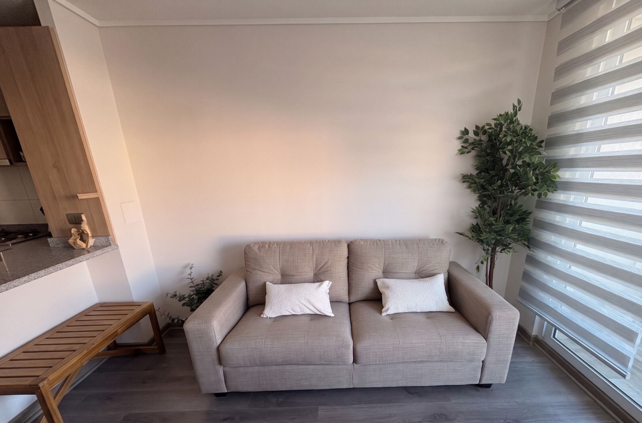 A living room with a beige sofa, a wooden bench, a potted plant, and a window with blinds.